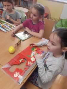 Trzy dziewczynki z oddziału przedszkolnego siedzą przy wspólnym stole. Przed każdą z nich otwarta jest książka o dinozaurach. Dziewczynki patrzą przed siebie.