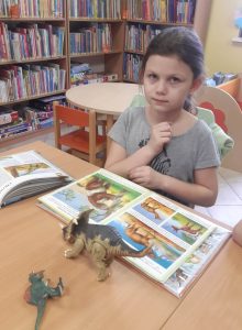 Uczennica oddziału przedszkolnego siedzi przy stoliku. Przed nią leży otwarta książka z rysunkami dinozaurów oraz dwa gumowe modele dinozaurów.
