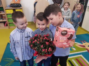 Trzech uczniów oddziału przedszkolnego pozuje do wspólnego zdjęcia. Chłopiec stojący po środku trzyma bukiet czerwonych kwiatów a chłopiec stojący po jego prawej stronie pochyla się w jego stronę trzymając w dłoniach pudełko z ptasim pleczkiem.