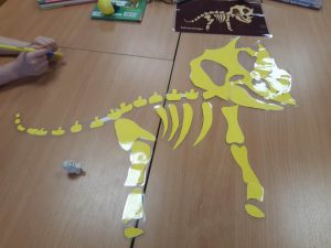 Zdjęcie rozłożonego na płasko na stole, żółtego szkieletu dinozaura. Szkielet wykonany z zalaminowanego papieru.