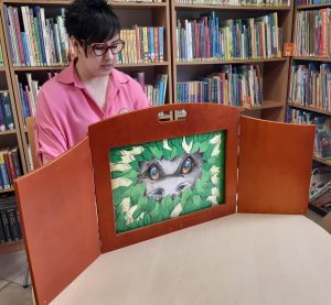 Pani bibliotekarka siedzi kontuarem imitującym scenę teatralną. Po środku widoczny jest rysunek oczu i ona zwierzęcia schowanego za liściami.