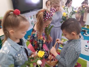 Uczeń oddziału przedszkolnego stoi na przeciwko ustawionych w rzędzie dziewczynek. Chłopiec trzyma w dłoniach koszyczek, a dziewczynka stojąca naprzeciwko niego wyciąga z koszyczka słodki upominek.