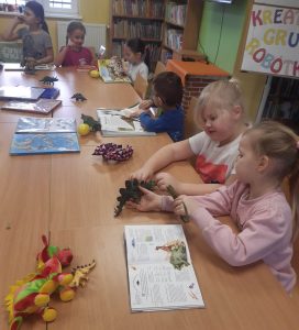 Uczniowie oddziału przedszkolnego siedzą przy długim stole w bibliotece w Ostaszewie. Na stole leżą porozkładane książki i modele oraz zabawki w kształcie dinozaurów. Dwie dziewczynki siedzące na pierwszym planie trzymają w dłoniach modele dinozaurów, którym przyglądają się z zaciekawieniem.