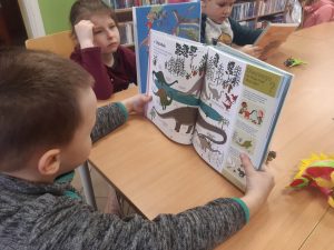 Troje uczniów oddziału przedszkolnego siedzi przy wspólnym stole. Każde z nich trzyma w dłoniach otwartą książkę z obrazkami przedstawiającymi dinozaury.