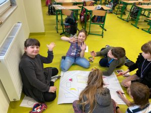 Uczniowie podczas wykonywania pracy plastycznej. Dzieci siedzą na podłodze a przed nimi leży duży arkusz papieru z narysowanym zarysem domu. Dzieci kolorują dom kredkami i pisakami. Dwie dziewczynki siedzące przodem do zdjęcia podnoszą w górę dłonie z szeroko rozstawionymi palcami.