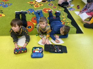 Trzech uczniów leży wyciągniętych wzdłuż na dywanie. Przed nimi leżą wycinki kół do pomalowania oraz piórniki.