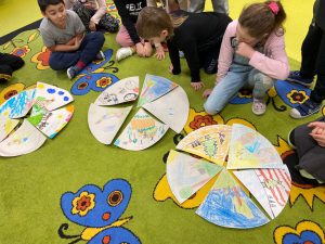 Uczniowie kucają na dywanie i przyglądają się leżącym na nim kołom złożonych wycinków papieru. Każdy wycinek został ręcznie pomalowany i przedstawia inny fragment omawianej książki.