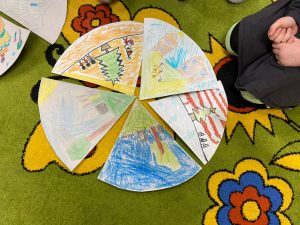 Na podłodze ułożony jest obrazek z pięciu wycinków tworzących koło. Każdy wycinek przedstawia inny obrazek wykonany przez uczniów kredkami.