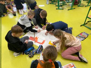 Uczniowie podczas wykonywania pracy plastycznej. Dzieci siedzą na podłodze a przed nimi leży duży arkusz papieru z narysowanym zarysem domu. Dzieci kolorują dom kredkami i pisakami.