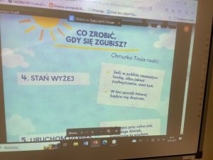 Zdjęcie monitora na którym wyświetlony jest slajd prezentacji. Na niebieskim tle z grafiką słońca oraz chmur napisy: "CO ZROBIĆ, GDY SIĘ ZGUBISZ?" Chmurka Tosia radzi: Jeśli w pobliżu zauważysz ławkę, albo jakieś podwyższenie, stań tam. W ten sposób łatwiej będzie cię dostrzec." Po lewej stronie od napisy tekst: "4. STAŃ WYŻEJ".