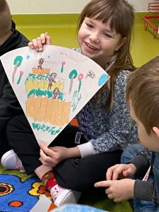 Dziewczynka siedzi w siadzie skrzyżnym na podłodze. Uśmiecha się. W dłoniach przed sobą trzyma wycinek koła, na którym narysowała scenę z omawianej książki.