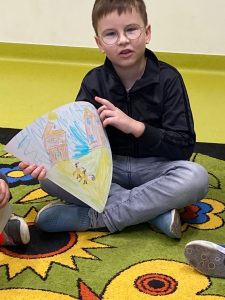 Chłopiec siedzi w siadzie skrzyżnym na podłodze. W dłoniach przed sobą trzyma wycinek koła, na którym narysowała scenę z omawianej książki.