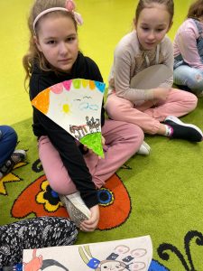 Dwie dziewczynki siedzą w siadzie skrzyżnym na dywanie. Jedna z nich trzyma przed sobą wycinek koła, na którym namalowała scenkę z omawianej książki.