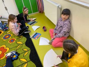 Cztery dziewczynki siedzą na podłodze. Przed nimi leżą wycinki kół przygotowane do stworzenia prac plastycznych. Na podłodze leżą również pisaki. Jedna z dziewczynek opiera się plecami o ściane i spogląda w bok.