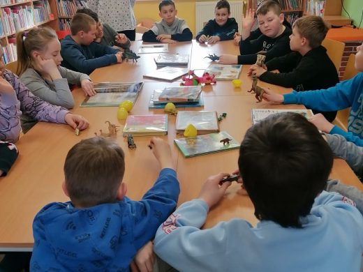 Uczniowie klasy trzeciej siedzą przy długim stole. Dzieci oglądają książki i modele dinozaurów ustawione na stole.