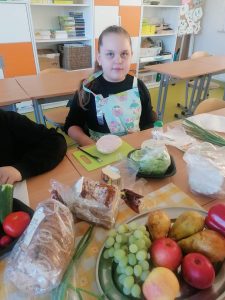 Uczennica klasy trzeciej podczas wykonywania kanapki. Dziewczynka siedzi przy stoliku na którym leży pieczywo, warzywa i owoce.