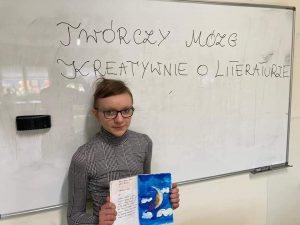 Uczennica klasy czwartej stoi przy tablicy z napisem "TWÓRCZY MÓZG KREATYWNIE O LITERATURZE". Dziewczynka trzyma przed sobą otwarty zeszyt z rysunkiem księżyca wśród chmur.