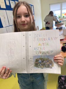 Uczennica klasy czwartej pozuje do zdjęcia z otwartym zeszyt, w którym widać odręczny, kolorowy napis: "DZIECI Z BULLERBYN AUTOR ASTRID ZIINGREN". Pod spodem naklejony jest obrazek przedstawiający gromadkę dzieci.