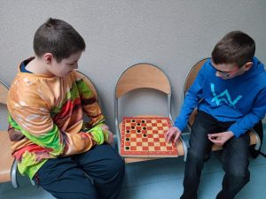 Dwóch uczniów klasy siódmej podczas gry w warcaby. Chłopcy siedzą na krzesełkach na szkolnym korytarzu. Szachownica leży rozstawiona na krześle pomiędzy nimi.