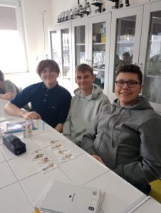 Trzech uczniów klasy ósmej uśmiecha się do zdjęcia. Na białym stole przy którym siedzą widać rozłożone karty do gry.