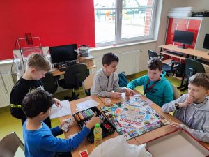 Uczniowie klasy czwartej podczas gry w monopoly. Pięciu chłopców siedzi przy stole na którym rozłożona jest gra.