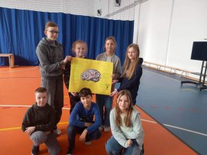 Uczniowie klasy czwartej pozują do wspólnego zdjęcia z wykonanym plakatem na którym widać przekreśloną grafikę mózgu. Czworo uczniów trzyma plakat a troje pod nim kuca.