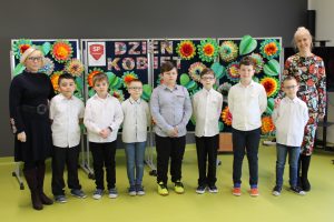 Uczniowie w białych koszulach pozują do wspólnego zdjęcia z nauczycielką języka polskiego i Panią Dyrektor. Zdjęcie wykonane na sali teatralnej. W tle za chłopcami stoi tablica z kolorowym napisem "Dzień Kobiet" oraz dużymi kolorowymi kwiatami i zielonymi liściami uformowanymi z papieru.