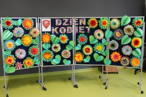Zdjęcie tablicy przygotowanej z okazji Dnia kobiet. Na środku widnieje kolorowy napis "Dzień Kobiet" a wokół niego umieszczono duże kolorowe kwiaty i zielone liście uformowane z papieru.