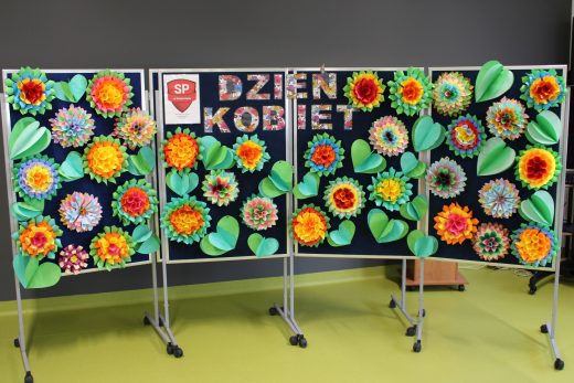 Zdjęcie tablicy przygotowanej z okazji Dnia kobiet. Na środku widnieje kolorowy napis "Dzień Kobiet" a wokół niego umieszczono duże kolorowe kwiaty i zielone liście uformowane z papieru.