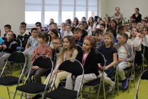 Uczniowie szkoły siedzą na krzesełkach na sali teatralnej. W tle widać stojące dwie nauczycielki.