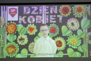 Zdjęcie wykonane podczas wyświetlania filmu. Na tle z kolorowymi kwiatami i napisem "Dzień Kobiet" stoi uczeń w białej koszule. U doły wyświetlony jest napis: "Zanim wiosna nastanie, jest czas na świętowanie."