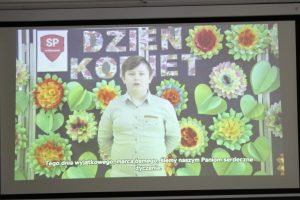 Zdjęcie wykonane podczas wyświetlania filmu. Na tle z kolorowymi kwiatami i napisem "Dzień Kobiet" stoi uczeń w białej koszule. U doły wyświetlony jest napis: "Tego dnia wyjątkowego, marca ósmego, ślemy naszym Paniom serdeczne życzenia."