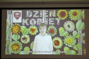 Zdjęcie wykonane podczas wyświetlania filmu. Na tle z kolorowymi kwiatami i napisem "Dzień Kobiet" stoi uczeń w białej koszule. U doły wyświetlony jest napis: "Bardzo Was wszystkie doceniamy i całusy przesyłamy."