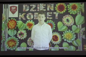 Zdjęcie wykonane podczas wyświetlania filmu. Na tle z kolorowymi kwiatami i napisem "Dzień Kobiet" stoi uczeń w białej koszule. U doły wyświetlony jest napis: "to z Wami każdego dnia to święto celebrować będziemy."