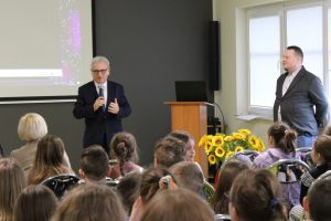 Starosta toruński stoi z mikrofonem i przemawia do siedzących naprzeciwko uczniów. Pod oknem stoi Pan w garniturze.