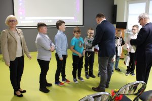 Uczniowie występujący w filmie stoją w jednym rzędzie na sali teatralnej. Za nimi stoi uśmiechnięta Pani Dyrektor. Na przeciwko uczniów stoi dwóch Panów rozdających chłopcom upominki w białych papierowych torbach.