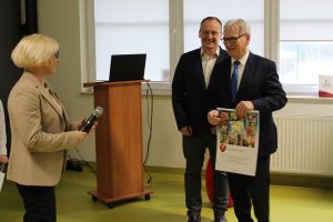 Pani Dyrektor stoi z mikrofonem na sali teatralnej. Na przeciwko niej stoi Pan Starosta trzymający otrzymany w prezencie od szkoły kalendarz. Za nim stoi uśmiechnięty Pan w czarnym garniturze.