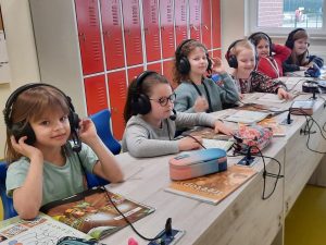 Sześć uczennic klasy pierwszej siedzi przy długim stole. Dziewczynki mają założone na uszach słuchawki z mikrofonami podłączone kablem do stołu. Przed nimi leżą książki i piórniki.