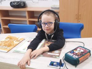 Uczeń klasy pierwszej siedzi przy stole ze słuchawkami na głowie. Uczeń wyciąga daleko przed siebie ręce. Przed nim leży otwarta książka i piórnik.