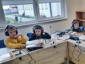 Trzech uczniów klasy pierwszej siedzi przy wspólnym stoliku. Chłopcy mają na uszach słuchawki z mikrofonami podłączone kablami do stołu. Przed nimi leżą książki i piórniki.