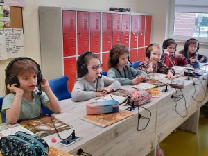 Sześć uczennic klasy pierwszej siedzi przy długim stole. Dziewczynki mają założone na uszach słuchawki z mikrofonami podłączone kablem do stołu. Przed nimi leżą książki i piórniki.