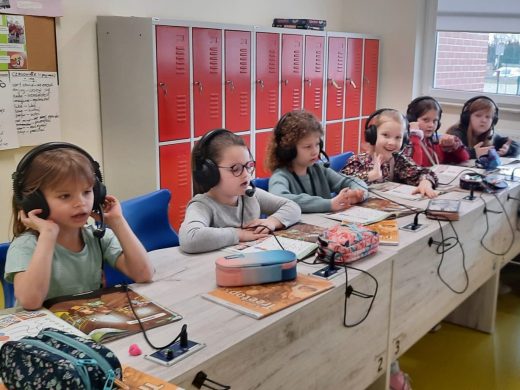 Sześć uczennic klasy pierwszej siedzi przy długim stole. Dziewczynki mają założone na uszach słuchawki z mikrofonami podłączone kablem do stołu. Przed nimi leżą książki i piórniki.