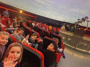 Uczniowie siedzą w czerwonych fotelach na sali w planetarium. Fotele ustawione są po okręgu. Część dzieci odwraca głowę w tył żeby byc widocznym na zdjęciu.