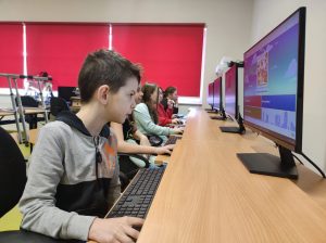 Uczniowie klasy czwartej siedzą w jednym rzędzie przy stanowiskach komputerowych. Uczniowie patrzą na ekrany umieszczone przed nimi.