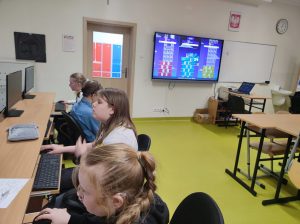Uczennice klasy czwartej siedzą obok siebie przy stanowiskach komputerowych. Jedna z uczennic jest odwrócona plecami do monitora. Na ścianie z boku widoczny jest monitor z wyświetlonym quizem.