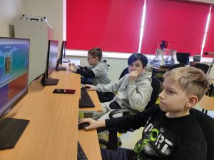 Trzech uczniów klasy czwartej siedzi obok siebie przy stanowiskach komputerowych. Dwóch uczniów patrzy na ekrany przed sobą a uczeń siedzący pomiędzy nimi spogląda w bok trzymając dłoń przy ustach.