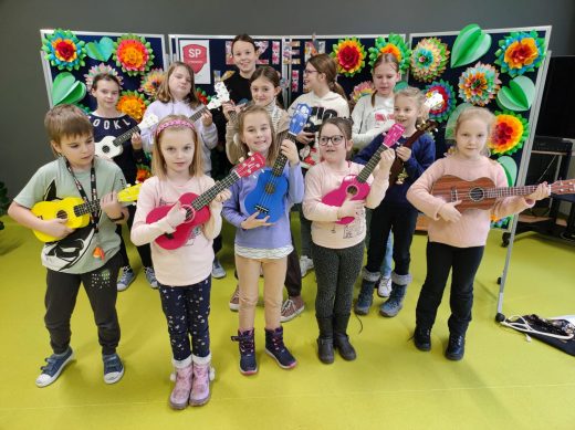 Uczniowie biorący udział w zajęciach gry na ukulele stoją ze swoimi instrumentami na tle tablicy z kolorowymi kwiatami.