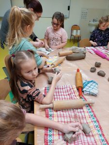 Uczniowie siedzą przy stoliku i wykonują figurki z gliny. Na stole leżą rozłożone kuchenne ręczniki, walki oraz kawałki gliny. Pomiędzy dwoma dziewczynkami stojącymi na końcu stołu stoi Pani pomagająca im w pracy.