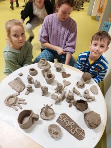 Dwoje uczniów klasy pierwszej wraz z wolontariuszką z klasy piątej siedzi przy okrągłym stoliku. Na stoliku ustawiono figurki wykonane przez uczniów podczas warsztatów.