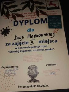 Zdjęcie dyplomu otrzymanego przez Łucję Markowską. Na białym tle ozdobionym u góry grafiką zielonych liści i logo gminy Łysomice, następujące napisy:" DYPLOM dla Łucji Markowskiej za zajęcie II miejsca w konkursie plastycznym "Mikołaj Kopernik - człowiek nauki". Pod napisami pieczątki i podpisy organizatorów i dyrektora Szkoły Podstawowej w Świerczynkach. Na środku u dołu grafika globusa umieszczonego na stelażu w kształcie kielicha a pod nim napis: "Świerczynki 31.03.2023r.".